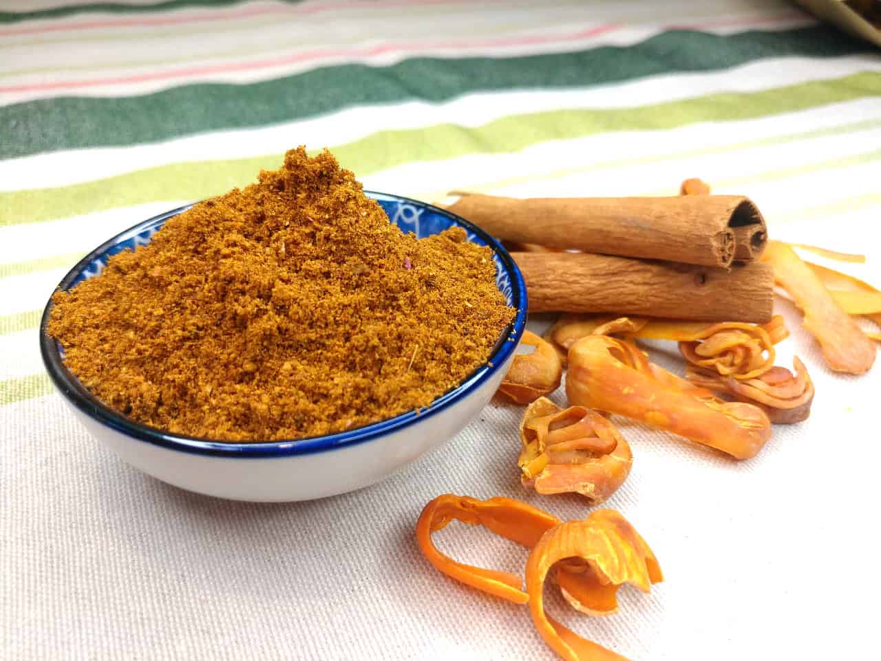 Homemade Ras El Hanout spice blend in a bowl, surrounded by cinnamon sticks, whole mace, and a brown paper spice bag.