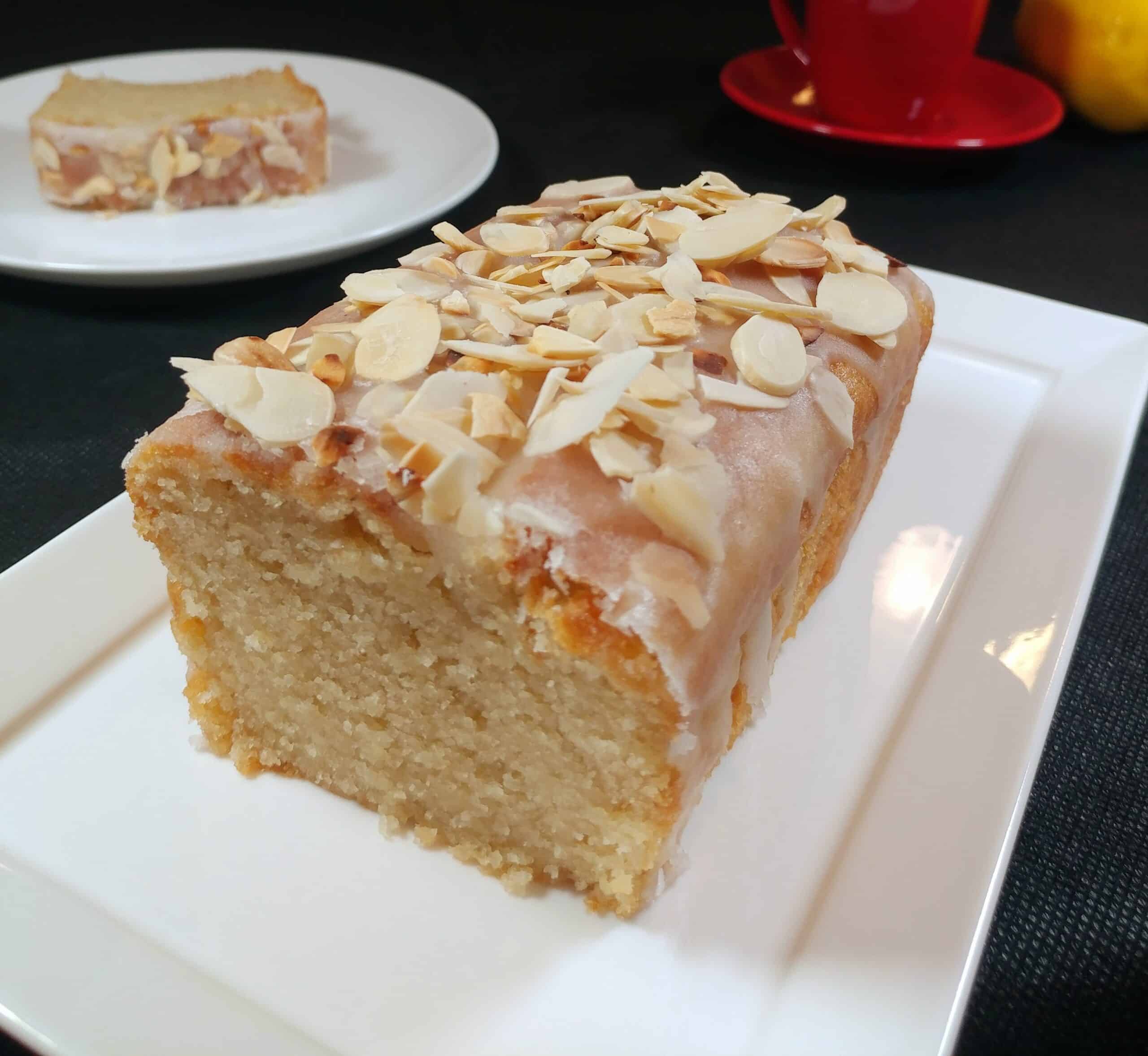 עוגת לימון טבעונית חתוכה לפרוסות על מגש מלבני לבן, עם פרוסת עוגה מוגשת על צלחת לבנה ברקע, מעל מפה שחורה.