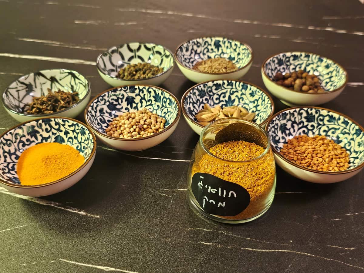 תערובת חוואיג' למרק בתוך צנצנת זכוכית, מוקפת בקעריות עם תבלינים, מונחת על משטח שיש שחור עם עורקים לבנים.