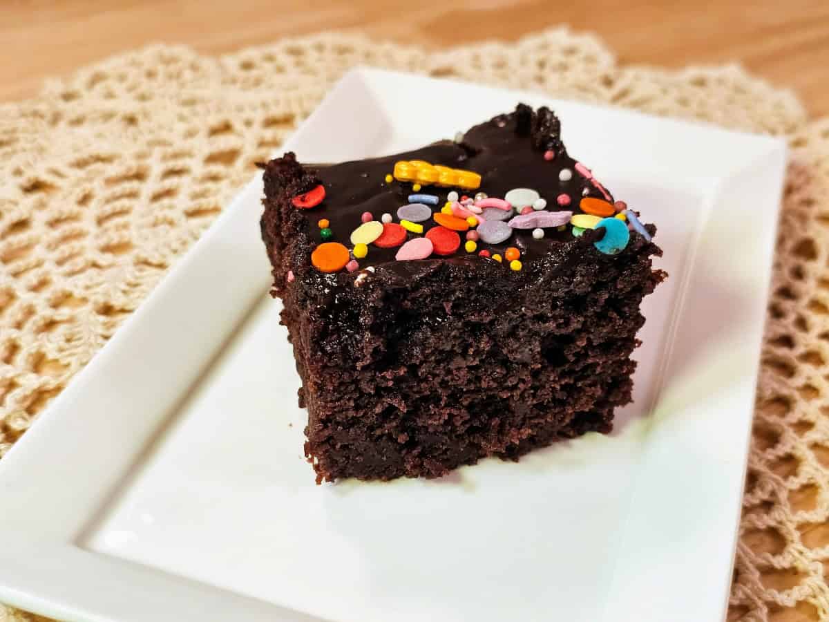 A slice of vegan chocolate cake on a white plate, topped with glossy chocolate glaze and sprinkles
